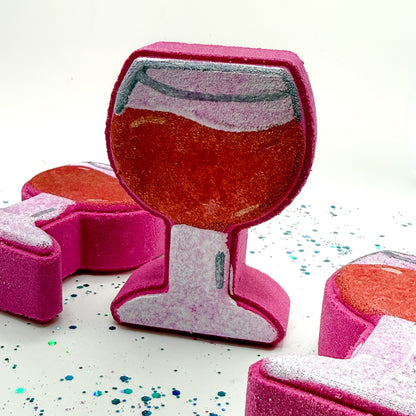 Three hand painted burgundy wine glass shaped bath bombs on a white background with blue and green glitter. Made by Rusted Acre Soap Co.