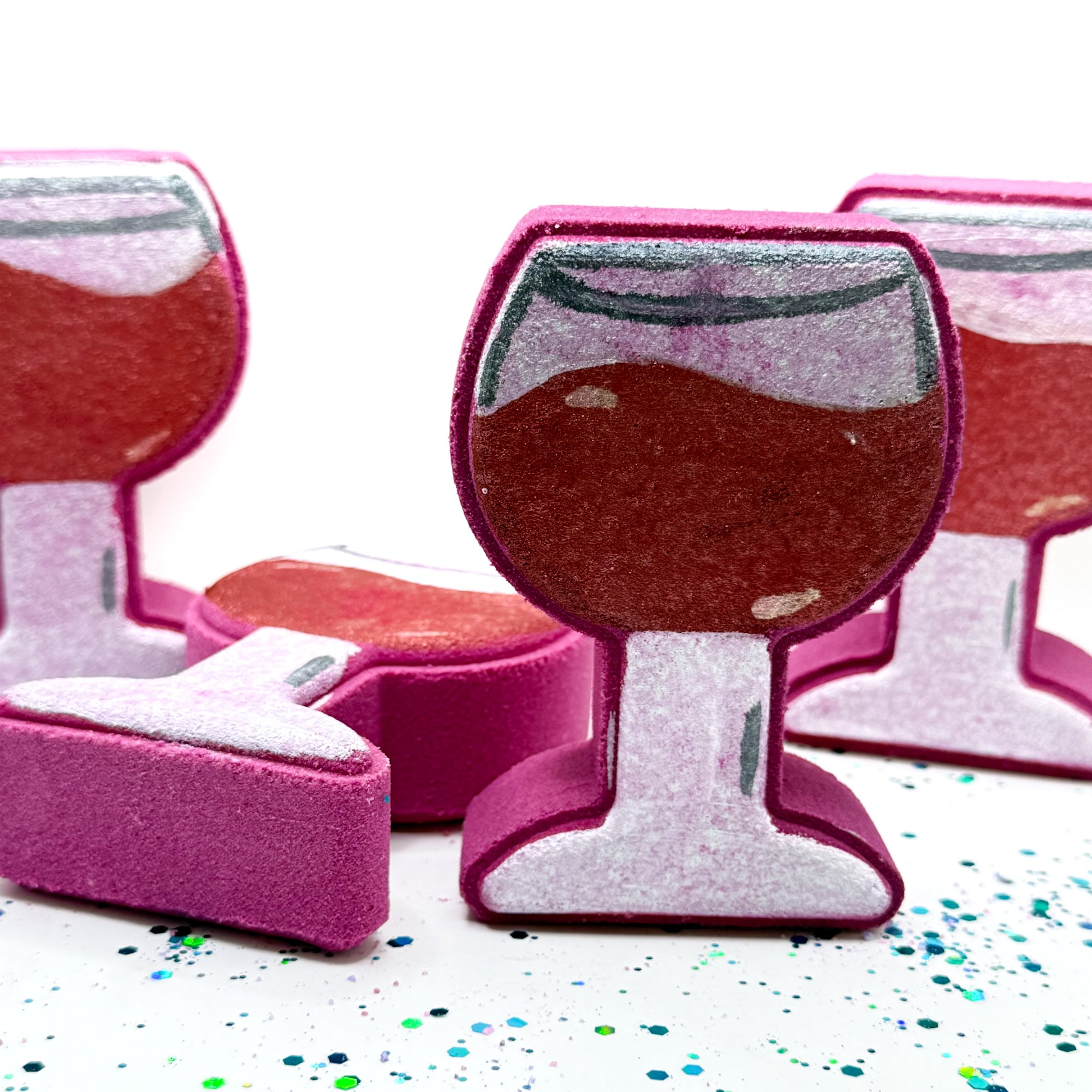 Multiple hand painted burgundy wine glass shaped bath bombs on a white background with blue and green glitter. Made by Rusted Acre Soap Co.