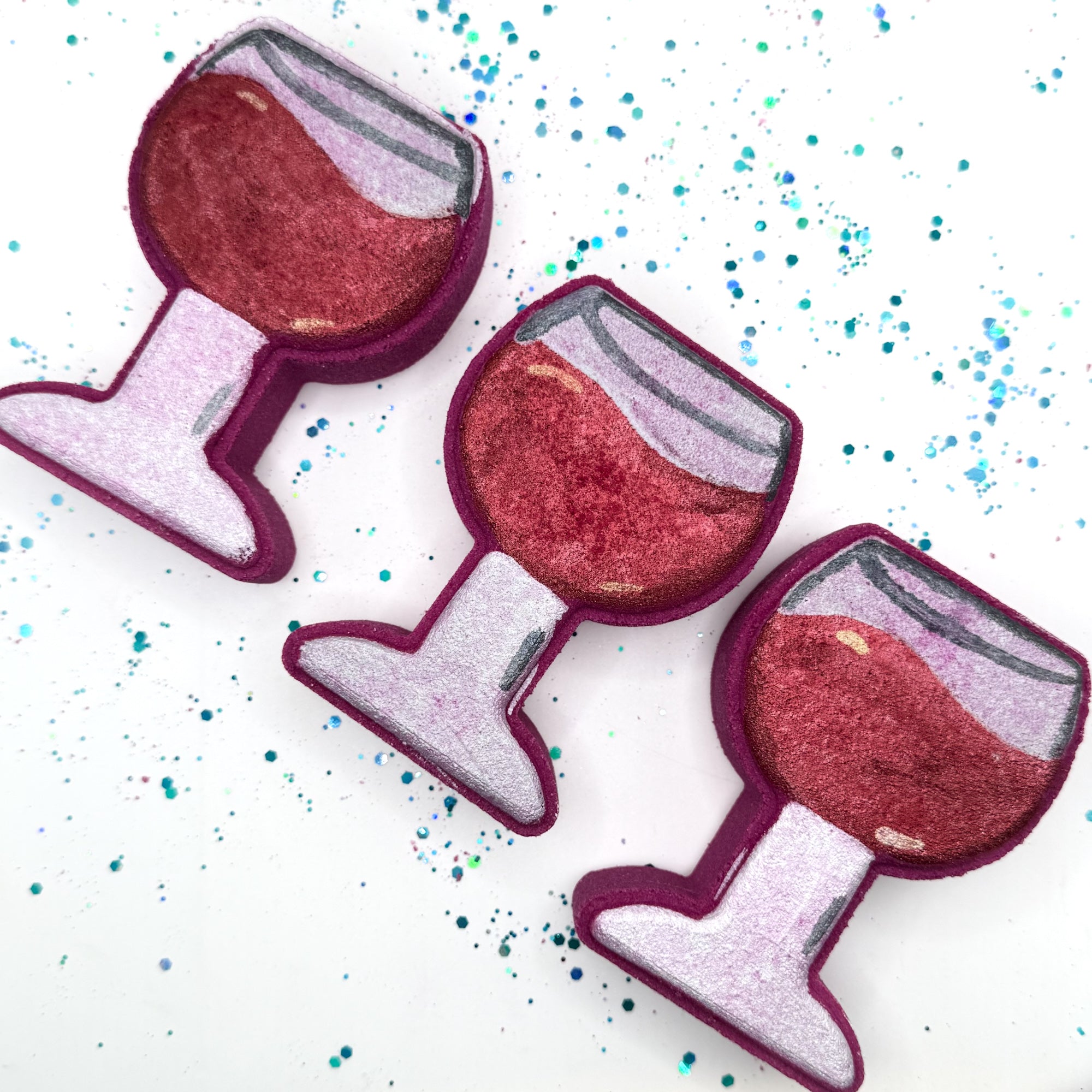 Three burgundy hand painted wine glass shaped bath bombs on a white background with blue and green glitter. 