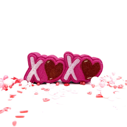 Pink bath bomb with raised white X's and pink hearts. Displayed with pink sprinkles of various shapes.